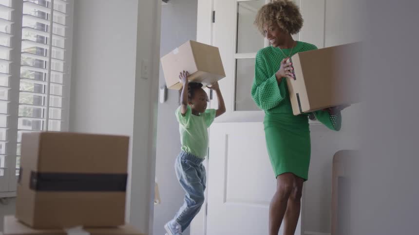 A smiling woman with blonde curls wearing a bright green outfit holds a large cardboard box while two small childs also carrying a box on their head walks past them into a sunny white entryway filled with moving boxes