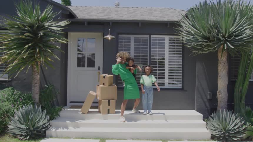 Woman standing on her porch next to a stack of boxes with her two children waving at the camera