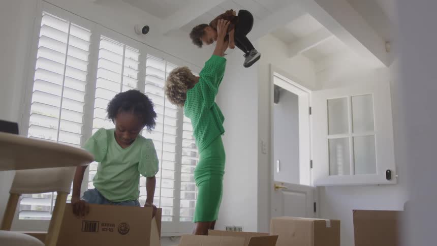 Mother plays with her son by lifting him into the air while her daughter plays with a cardboard box in their new home