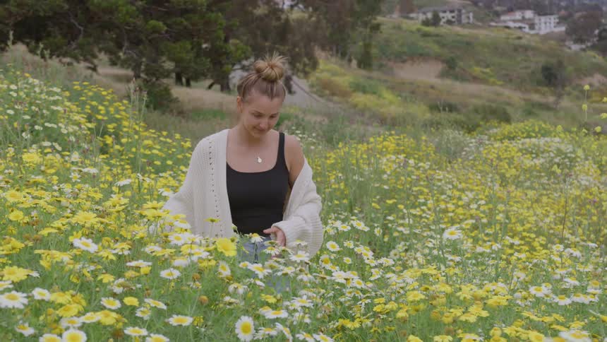 A woman wearing a black tank top and a light cardigan walks through a vibrant field abundant with yellow and white daisies she smiles while admiring the flowers in a hilly semiurban landscape