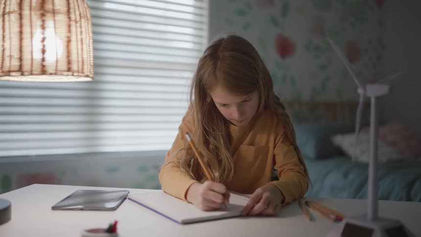 A young girl with long light hair leans over a white desk intently writing in a notebook with a pencil under the light of a hanging wicker lamp with a miniature wind turbine model and a tablet nearby
