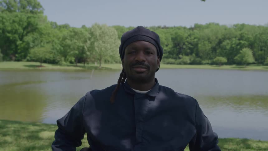 A man in a dark jacket and beanie sits outdoors by a lake on a sunny day turning his gaze to look straight at the camera he maintains steady eye contact with a calm neutral expression