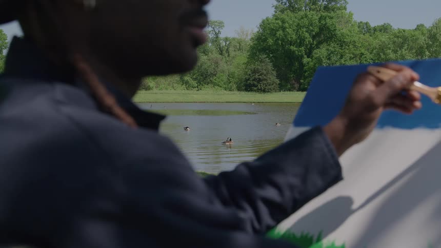 A painter is shown in a closeup overtheshoulder view applying blue and green paint to a canvas outdoors on a sunny day in the background a calm lake is visible with trees lining the shore and a few ducks swimming on the water