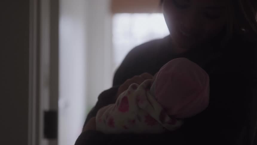 A caring adult mostly in silhouette stands indoors and tenderly cradles a newborn baby wrapped in a patterned blanket and pink cap with their faces partially obscured by darkness against a bright background light