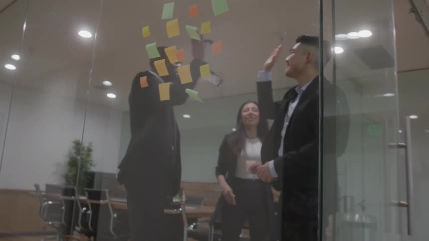 A camera pans up a hallway to a glasswalled office where two men and a woman highfive celebrating by a wall of sticky notes