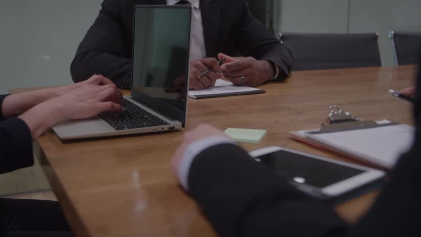 A closeup shot of a business meeting shows people in suits at a table with one person typing on a laptop and others holding pens