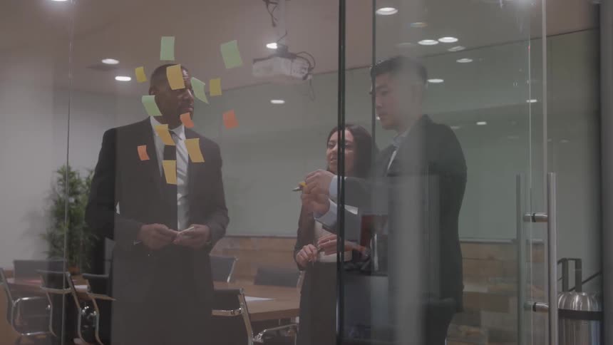 Three people standing in an office talking as a man sticks notes on a clear glass cubicle