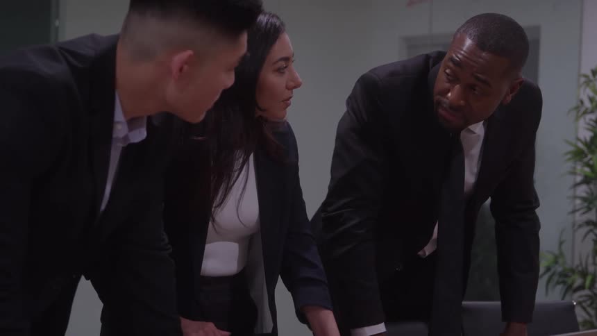 Three business professionals in black suits huddle around a table actively engaged in a discussion or reviewing material the man on the right is speaking and gesturing with his hand as the other two listen attentively