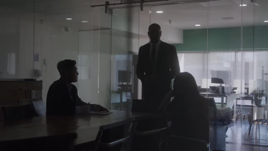 Two people are sitting at a table and a person standing is talking to them in an office meeting