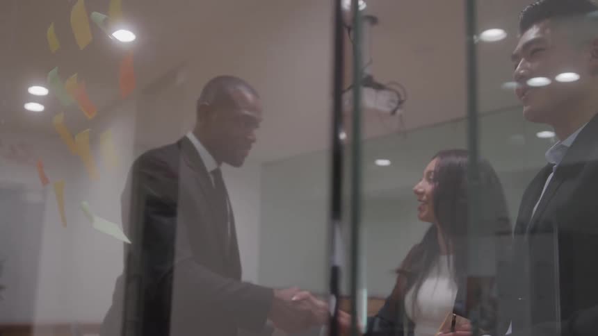 As seen through a glass wall in an office setting three people two of them are shaking hands while the third person looks on and smiles