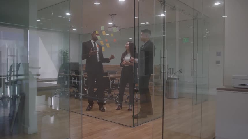 Three people standing in a room with clear glass cubicles having a meeting