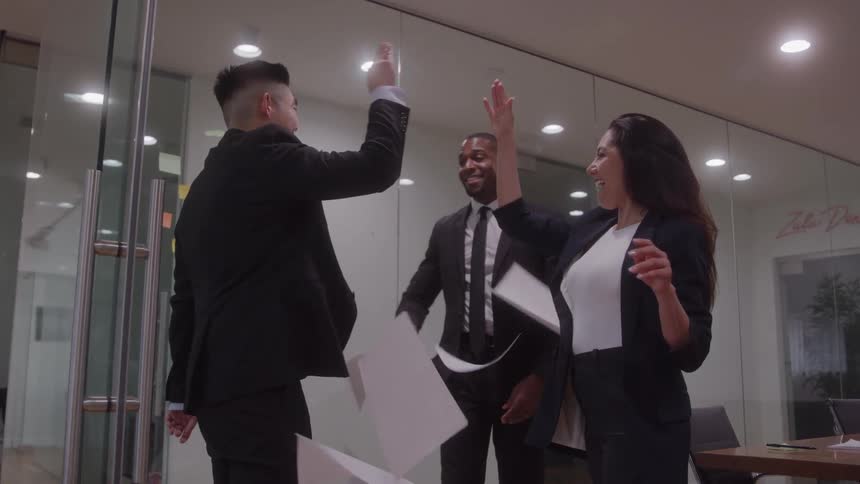 Three colleagues a black man an asian man and a white woman all wearing suits in an office lobby toss documents into the air and highfive each other in excitement