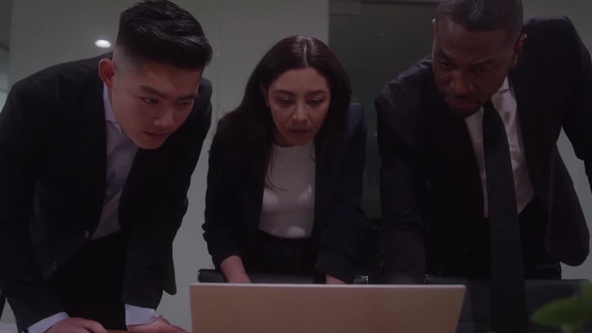 Three sharply dressed focused business professionals lean intently over a laptop computer placed on a dark conference table scrutinizing the screen together