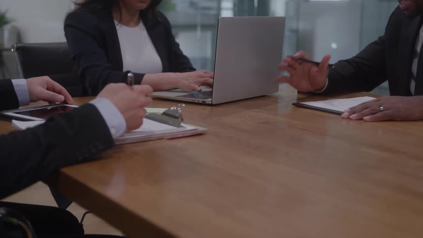 Three people sitting at an office table as the woman types on the laptop computer the men takes notes on paper