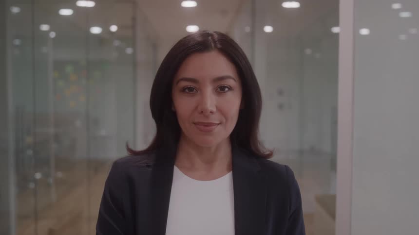 A professional woman with long dark hair and a confident expression stands still and looks directly at the camera framed by a modern brightly lit corporate office hallway featuring blurred glass walls and subtle background reflections
