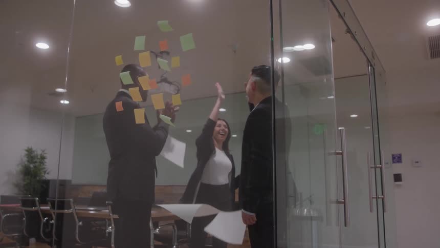 Three diverse business professionals in formal attire standing within a glasswalled conference room covered in colorful sticky notes celebrate a success by happily throwing papers into the air and sharing a highfive