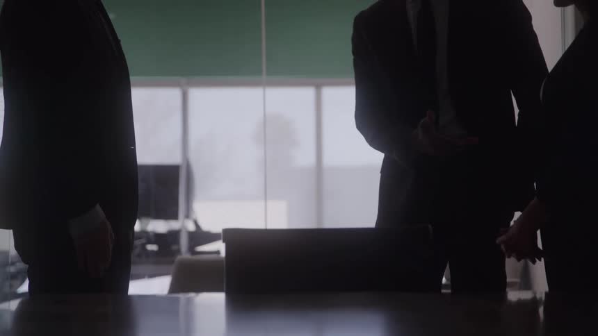In a dimly lit foreground three business professionals silhouetted against a bright large window shake hands over a conference table