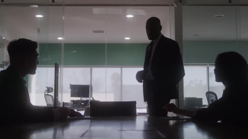 Two people are seated at a table listening to a male speaking standing nearby in a modern dimly lit office