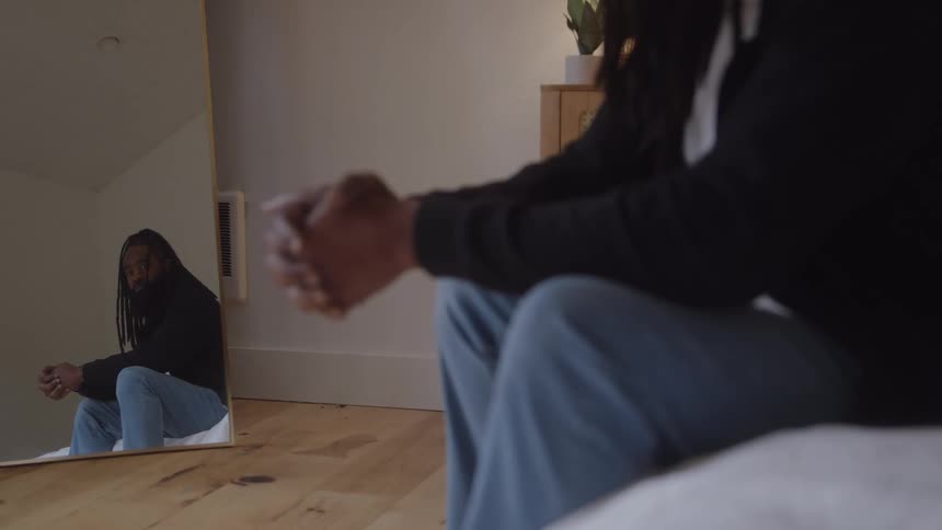 A man sits on a bed and looks at himself in a mirror