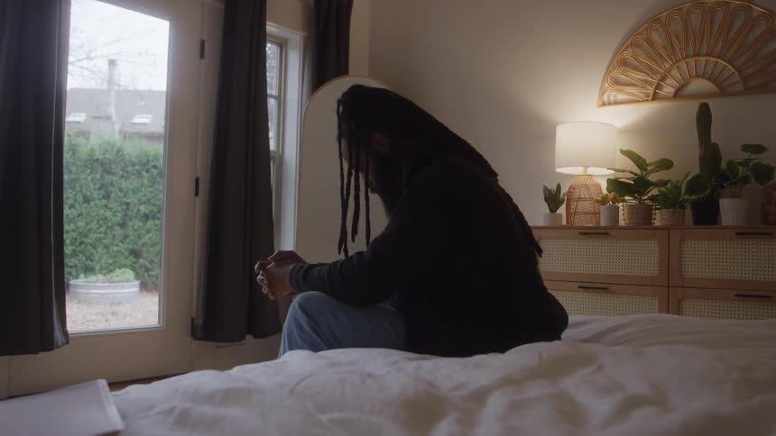 A black man sits on a bed with clasped hands and looking downward in deep thought