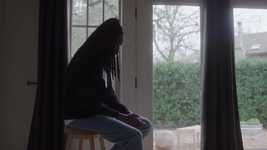 A man sitting on a bench next to a window is shaking his leg while holding his hands