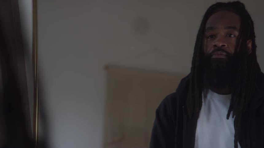 A man with long dreadlocks and a dense dark beard wearing a black jacket over a white shirt slowly lifts his head from a downward gaze to look directly with a serious contemplative expression in a dimly lit indoor setting