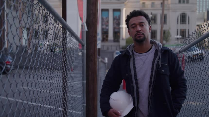 A man holding a white hard hat stands between two chainlink fences on a city street looking off before turning to the camera