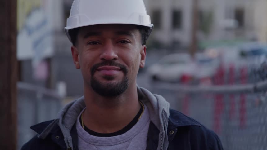 A focused male construction worker wearing a white hard hat over a dark jacket turns his head from looking offcamera to face forward breaking into a warm genuine smile while standing in front of a chain link fence at an urban job site