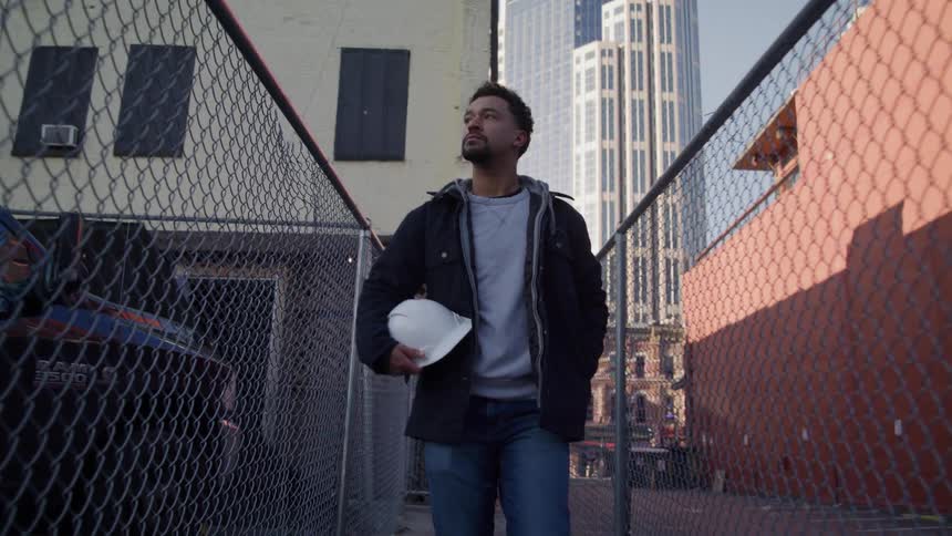 Man walks between two chain link fences near downtown while carrying a hard hat