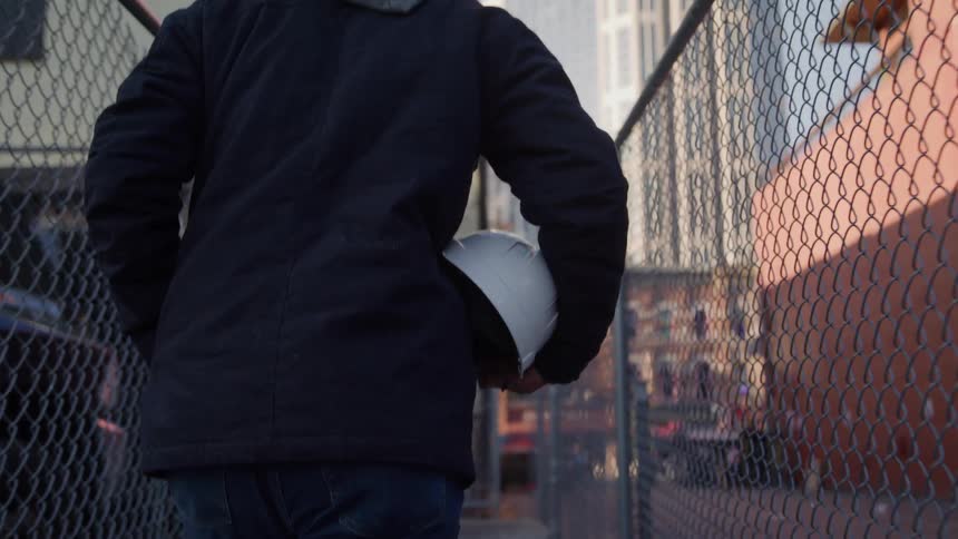 Man carrying a hard hat walks on the sidewalk between two fences in the city