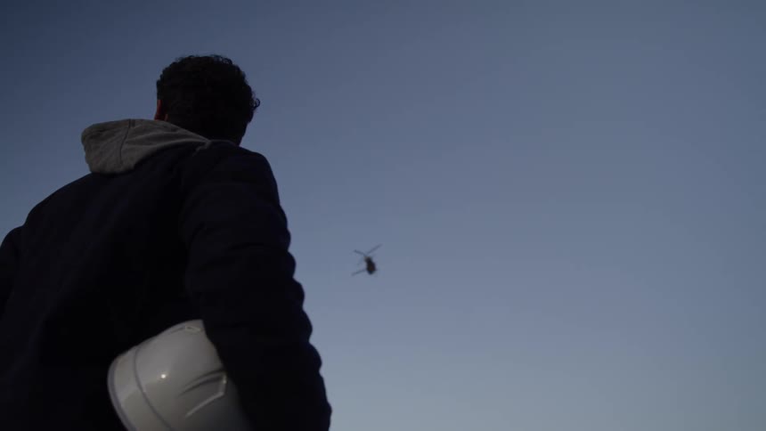 A lowangle shot from behind shows a man holding a hard hat and looking up as a helicopter flies across the blue sky