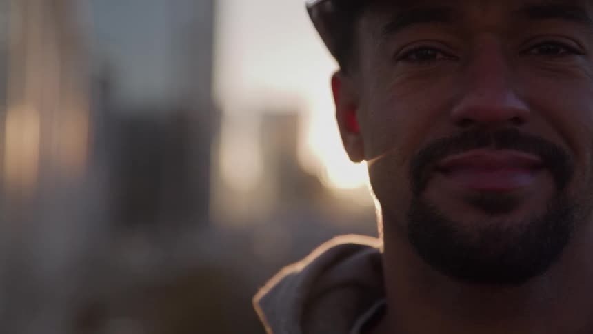 A tight closeup shows the right side of a smiling mans face wearing a hard hat and backlit by a bright sun flare
