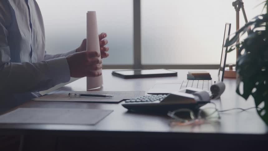 A person sits at a desk reviewing papers before picking up a pen to write on a document next to a calculator
