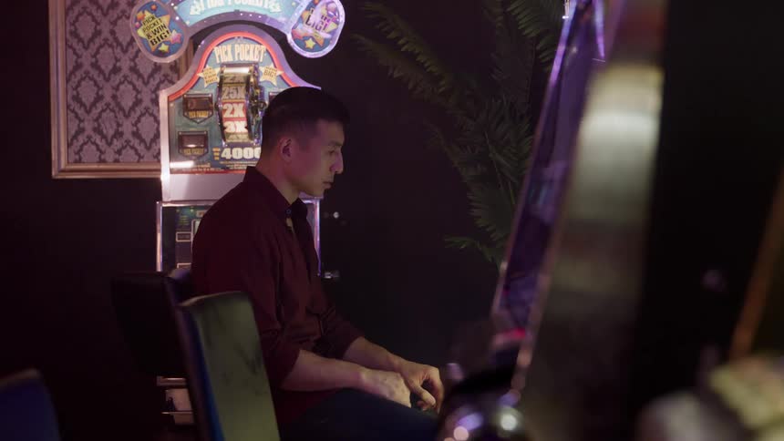 A man in a dark maroon shirt is seated leaning forward as he plays a brightly lit slot machine in a dimly lit indoor setting he pushes a button or lever and then glances upward seemingly watching the outcome of the game