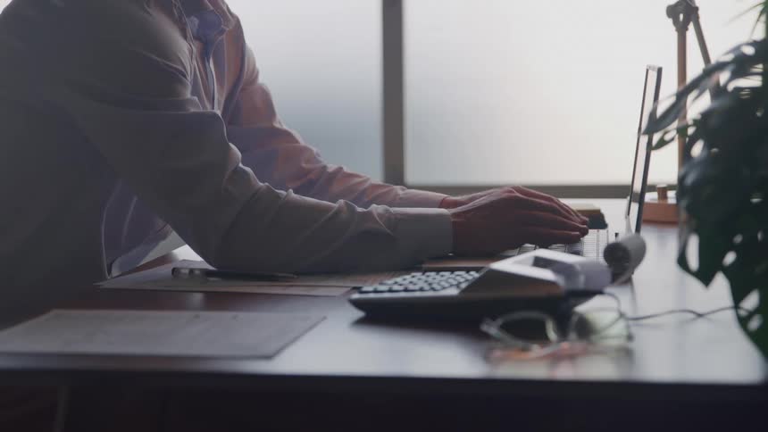 A person sits at a desk and types on a laptop with a calculator papers and eyeglasses on the desk nearby