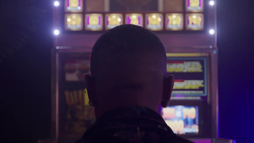 The back of a persons head is shown in a closeup dark silhouette as they stand in front of a brightly lit casino slot machine the machine flashes with various lights and symbols in the dark smoky environment