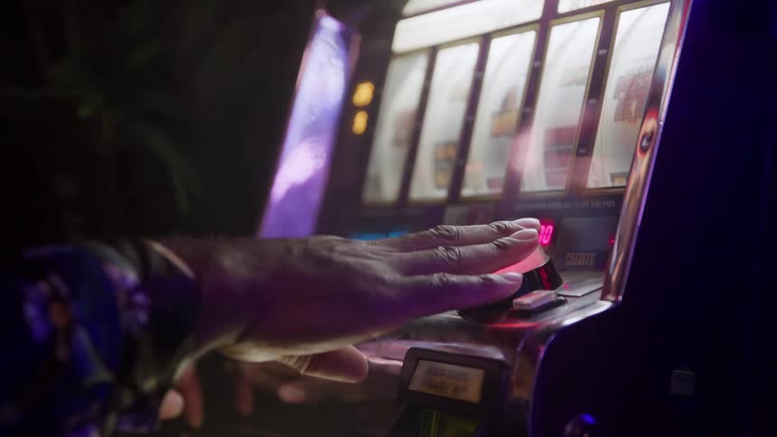 A closeup shot shows a persons hand pressing a red button to play a slot machine in a dark room