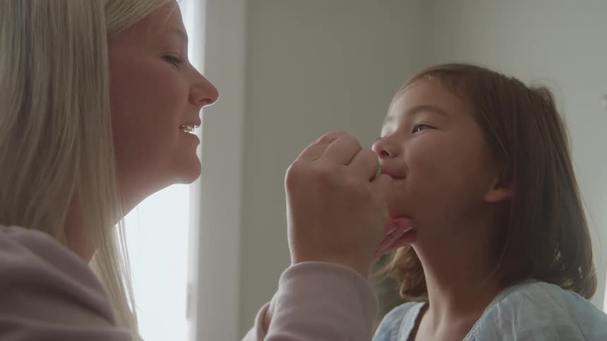 A mother applying lipstick to her daughters lips indoors at midday