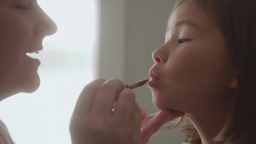 An adult woman is applying lip stick to the lips of a little girl who is looking at her