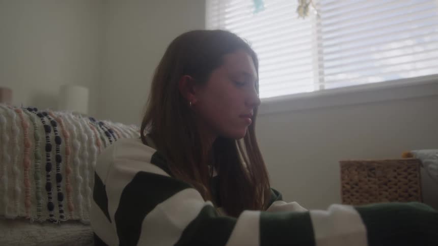 A young woman with long brown hair wearing a striped sweater is sitting alone on the floor of a sunlit bedroom next to a bed pulling her knees up and touching her hair before looking down with a somber expression