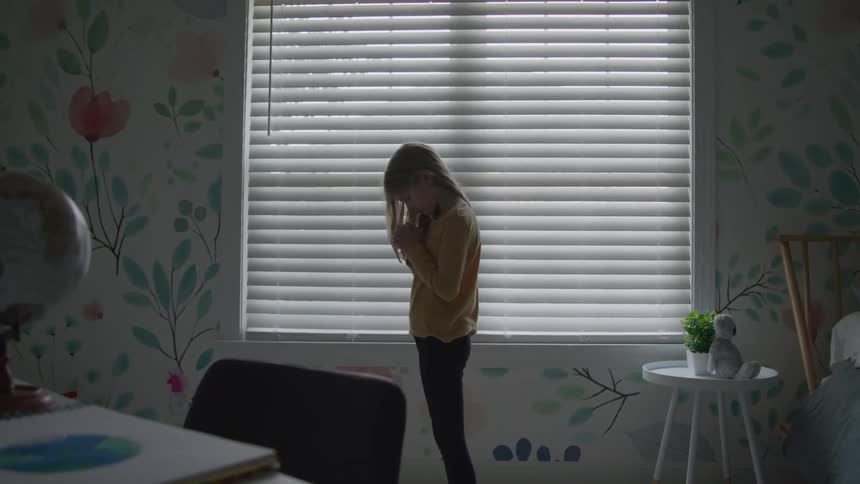 A young girl stands alone by a window in a softly lit bedroom