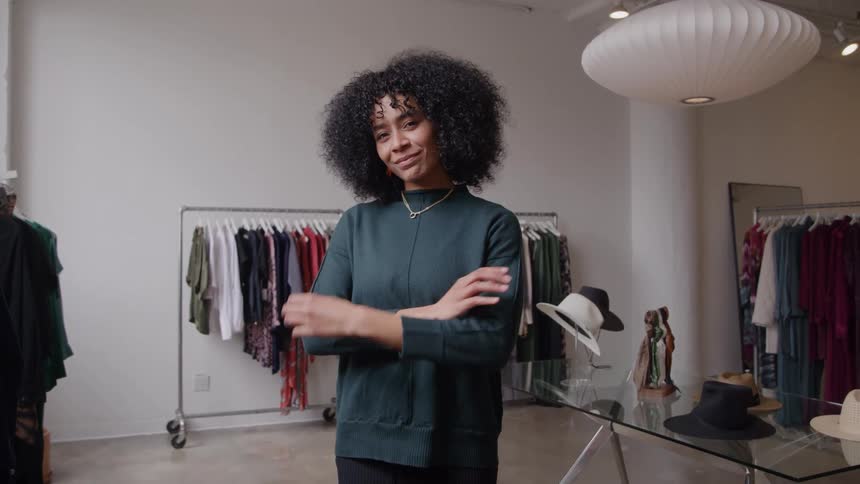 A woman with curly hair stands in a welllit clothing boutique initially with her eyes closed before opening them to smile confidently she folds her arms across her chest while standing among racks of clothing and a display of hats