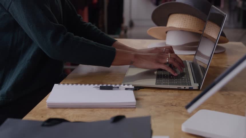 A person wearing a dark top diligently types on a silver laptop on a wooden counter with a notebook and pen resting nearby while behind her are various hats on display