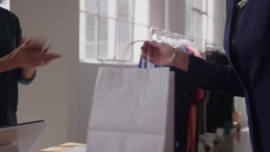 A customer dressed in a navy blazer is shown receiving a white shopping bag from an employee at the counter of a clothing store the customer takes the bag and then waves goodbye as they complete their transaction