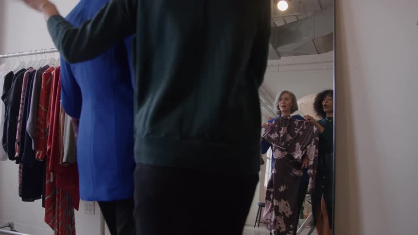 An older woman is helped by a younger one who holds up a dress in a hanger in front of the older woman as she checks how it would look on her on a mirror