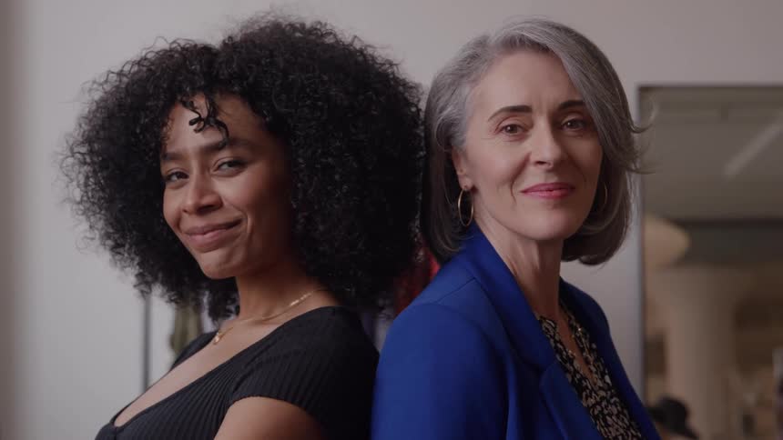 A younger woman and an older woman smile while standing back to back