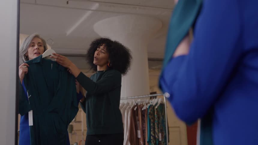 Two women one with grey hair wearing a blue blazer and another with dark curly hair wearing a dark green sweater are standing in a clothing boutique holding up a dark green dress to examine it in a large mirror