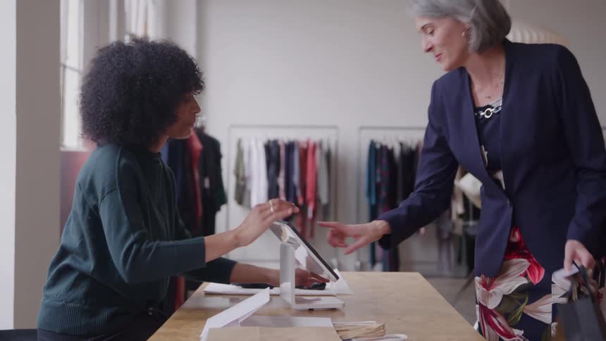 A young woman working at a dress shop and sitting at the payment counter talks and laughs with a standing client an older woman as the older woman types in her payment code on the digital terminal