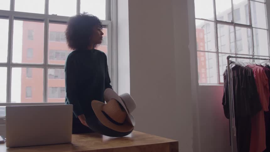 A young woman sitting at a payment desk in a clothes shop writes in a laptop then gets up and moves a pile of hats to a nearby table