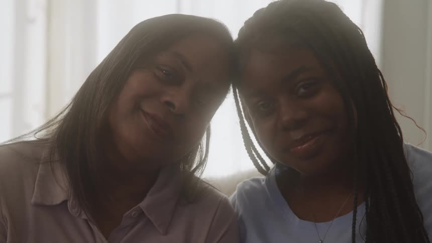 Two young black sisters sit indoors with smiles on their faces as they bump their heads together
